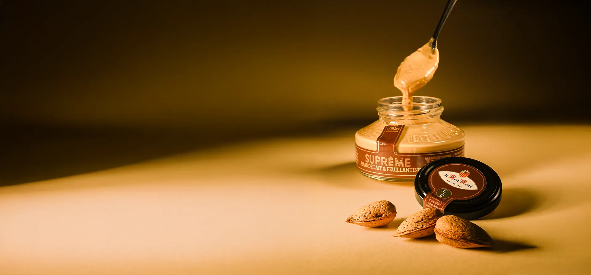 Un pot ouvert de pâte à tartiner amande lait et feuillantine du Roy René avec des amandes fraiches à coques posées à côté du pot. 