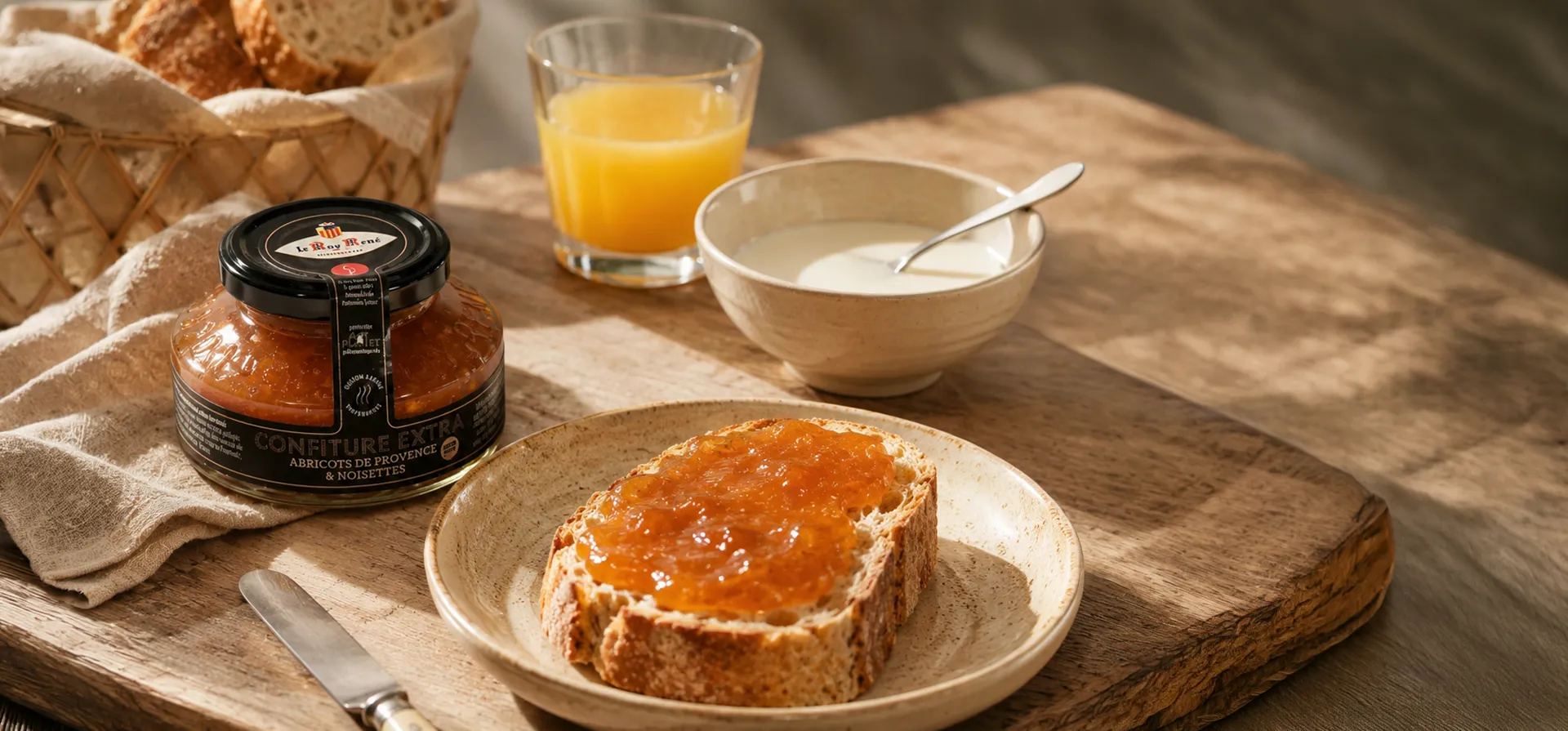 A jar of ‘Provence’ apricot jam by Roy René sitting on a table at breakfast, with a bowl of milk and a slice of toast with jam.